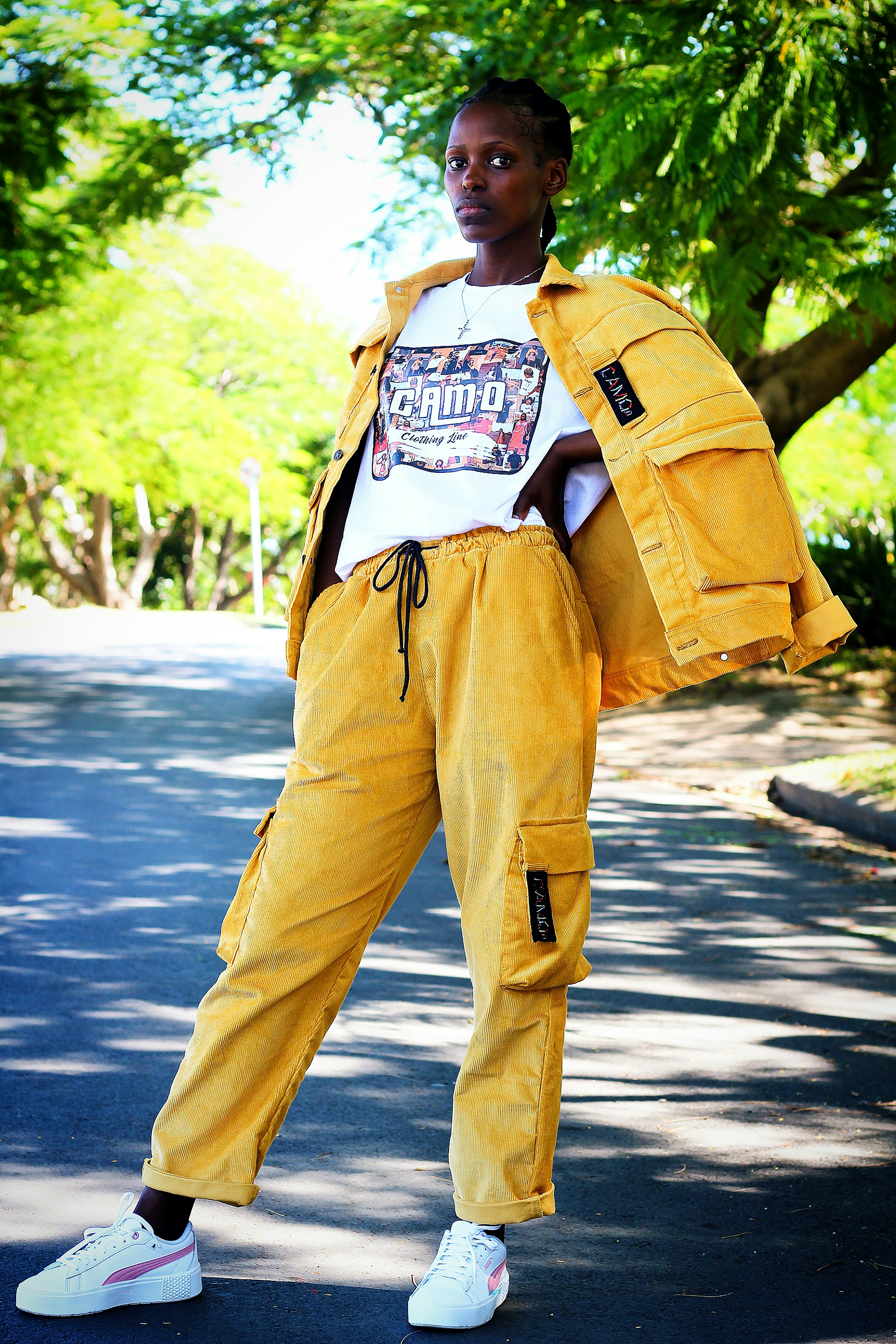 Woman in a yellow corduroy outfit posing on a tree-lined street.