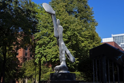 A smooth, abstract metal sculpture is prominently displayed in a park setting, surrounded by lush green trees under a clear blue sky. The sculpture's reflective surface catches light, creating bright highlights. Behind the sculpture, a red brick building and parts of a modern glass structure are visible.
