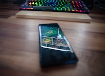 A smartphone displaying an image or webpage with a collage of different pictures lies on a wooden desk. Behind the phone, a colorful RGB backlit keyboard is visible, partially out of focus.