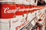 A row of stacked red and white soup cans on shelves, prominently displaying the brand name. The perspective creates a sense of depth, showcasing the canned products aligned perfectly in a supermarket aisle.