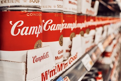 A row of stacked red and white soup cans on shelves, prominently displaying the brand name. The perspective creates a sense of depth, showcasing the canned products aligned perfectly in a supermarket aisle.