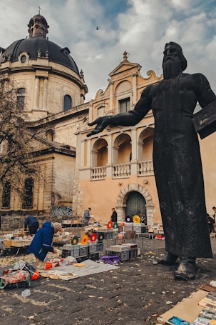 A market scene set outdoors in front of a historic building with an ornate dome and classical architecture. A large dark statue of a man is in the foreground, extending an arm outwards. Several people are browsing items laid out on the ground, including books and colorful vinyl records. The atmosphere suggests a casual market day.
