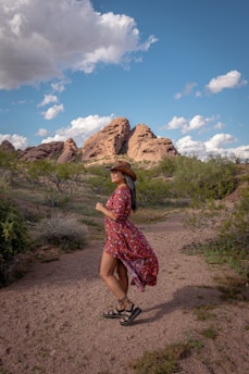 A person in a floral dress and cowboy hat strolls through a desert landscape. Rugged red rock formations stand prominently under a blue sky with scattered clouds. Sparse greenery dots the sandy terrain.