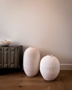 Two large, textured, white ceramic vases are placed on a wooden floor beside a dark, geometrically patterned cabinet. A small ornate vase sits on top of the cabinet, adding to the minimalist decor. The background is a plain, light-colored wall.