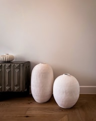 Two large, textured, white ceramic vases are placed on a wooden floor beside a dark, geometrically patterned cabinet. A small ornate vase sits on top of the cabinet, adding to the minimalist decor. The background is a plain, light-colored wall.