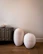 Two large, textured, white ceramic vases are placed on a wooden floor beside a dark, geometrically patterned cabinet. A small ornate vase sits on top of the cabinet, adding to the minimalist decor. The background is a plain, light-colored wall.