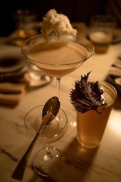 A set of three distinct drinks arranged on a table, with a cocktail glass containing a creamy beverage topped with a white foam, a smaller glass with a spoon inside, and a tall glass with a dark leaf garnish. The background is softly lit, with a few blurred objects seen in the distance, creating a warm and elegant ambiance.