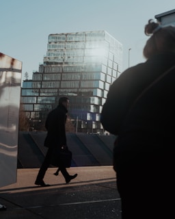 A modern office building with glass windows reflecting sunlight is in the background. A person is walking across the pavement in the foreground, carrying a briefcase. The image has a silhouette effect due to the backlighting.