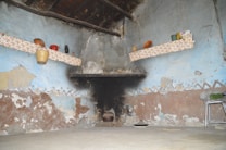The image features a rustic kitchen or storage area with a textured and aged wall that shows patches of peeling paint in shades of blue and brown. Two shelves lined with floral-patterned fabric hold a variety of items, including a basket, a red kettle, a lantern, and some containers and food items. The stone fireplace is prominent in the center, showing signs of soot and wear. The floor is simple and bare, with a metallic stool on the right side.
