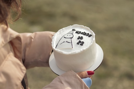 A small round white cake with smooth frosting and a simple cartoon-like drawing on top, held in someone's hands. The hands are wearing a light-colored coat and the nails are painted red. The cake features the text 'update to 22' along with the drawing.