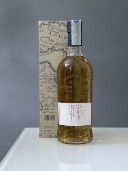 A bottle of Scotch whisky is placed in front of a box with a vintage map design. The label on the bottle includes text indicating it's from the Ardnamurchan Distillery in Scotland. The packaging and bottle are set on a plain white surface, with a grayish background.