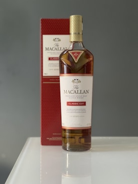 A bottle of Macallan Highland Single Malt Scotch Whisky labeled 'Classic Cut' is placed on a reflective white surface. The bottle is accompanied by its red and white box positioned in the background.
