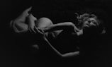 A black and white image featuring a nude person lying in a relaxed pose with closed eyes. The lighting casts soft shadows that emphasize the contours and details of the body, creating a serene and intimate atmosphere.