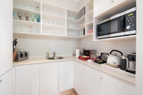 A compact kitchen area features white cabinets and open shelves filled with various glassware and cups. A countertop holds a sink, a cutting board with knives, a toaster, a kettle, a coffee machine, and a microwave. The decor is minimal, with a small vase of flowers adding a touch of elegance.