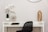 A minimalist workspace or decor setup featuring a white desk with a black chair. On the desk, there's a white pot with delicate white orchids, a decorative brass starburst sculpture, and a tall, hammered gold vase. Two books are neatly stacked underneath the starburst sculpture. The wall in the background is a soft, neutral color, and a round mirror is partly visible above the desk.