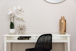 A minimalist workspace or decor setup featuring a white desk with a black chair. On the desk, there's a white pot with delicate white orchids, a decorative brass starburst sculpture, and a tall, hammered gold vase. Two books are neatly stacked underneath the starburst sculpture. The wall in the background is a soft, neutral color, and a round mirror is partly visible above the desk.