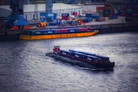 A port scene featuring a large barge carrying shipping containers on a river, with a smaller tugboat accompanying it. The background shows a dock area filled with stacked containers, cranes, and industrial buildings.