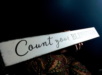 A person holds a long, rectangular sign with the words 'Count your BLESSINGS' written on it. The background is dark, and the person’s hands are visible, with a colorful patterned fabric beneath the sign.