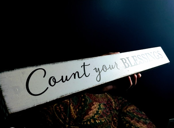 A person holds a long, rectangular sign with the words 'Count your BLESSINGS' written on it. The background is dark, and the person’s hands are visible, with a colorful patterned fabric beneath the sign.