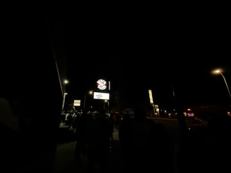 A dark outdoor scene featuring a neon sign with 'Space' written on it and an illuminated marquee underneath announcing a virtual event. Several street lamps light up the area, revealing silhouettes of people standing nearby. Additional signs are visible, including one indicating a motel.