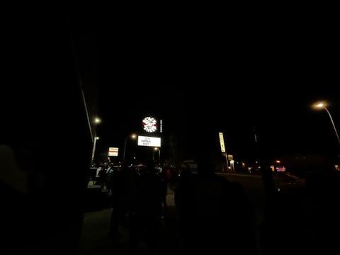 A dark outdoor scene featuring a neon sign with 'Space' written on it and an illuminated marquee underneath announcing a virtual event. Several street lamps light up the area, revealing silhouettes of people standing nearby. Additional signs are visible, including one indicating a motel.