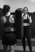 Two women are in a gym setting, with one holding a boxing pad while the other is positioned as if preparing to punch. Both are in athletic wear, focused and engaged in their activity. The environment appears to be a training room with padded flooring and walls.