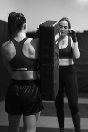 Two women are in a gym setting, with one holding a boxing pad while the other is positioned as if preparing to punch. Both are in athletic wear, focused and engaged in their activity. The environment appears to be a training room with padded flooring and walls.