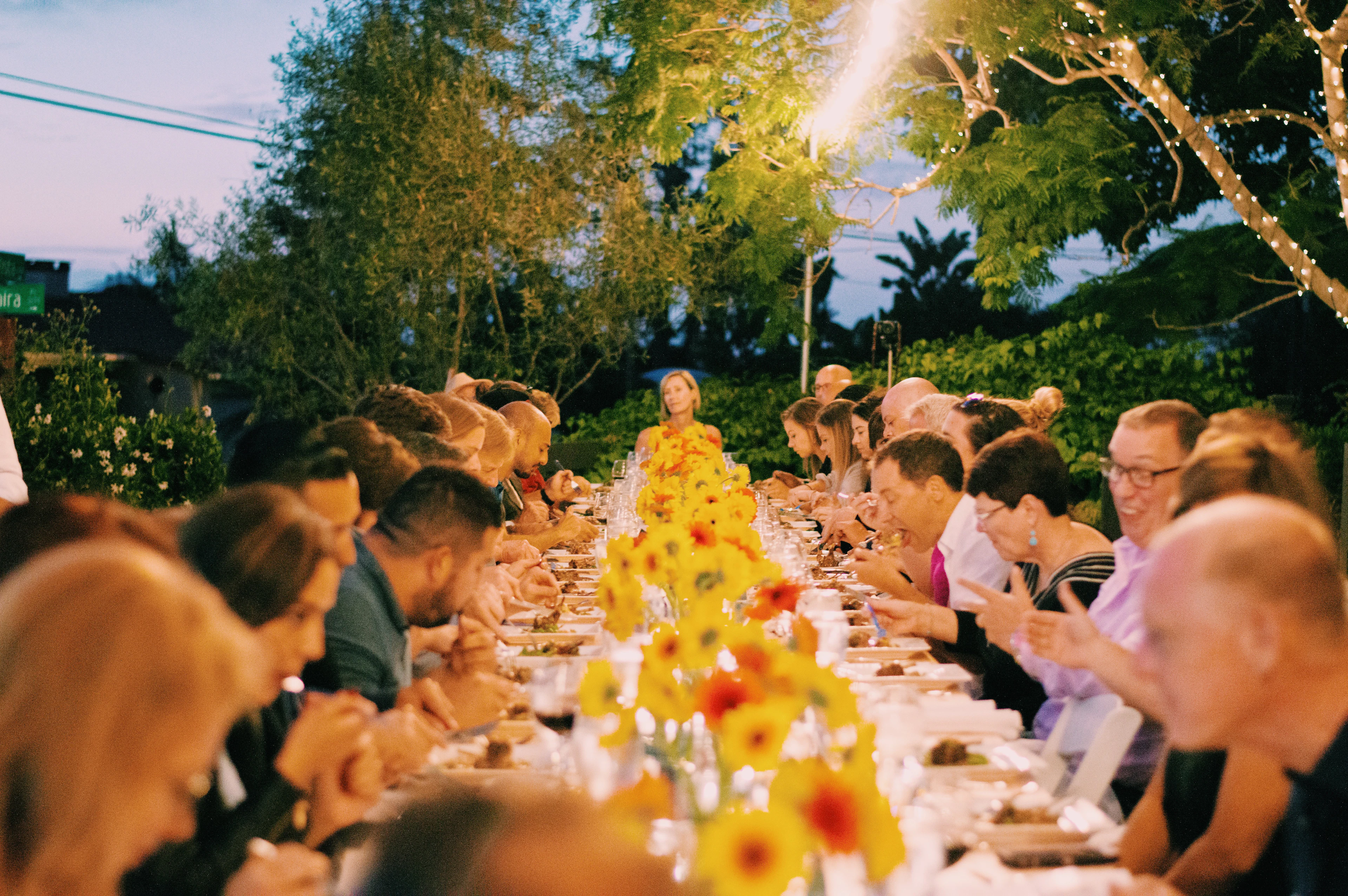Invités attablés le long d'une grande table fleurie de jaune, sous le soleil du soir