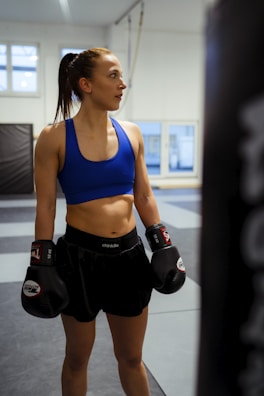 A female athlete in a gym setting wearing boxing gloves, a blue sports bra, and black shorts stands with an expression of focus and determination. The background features padded walls and large windows letting in natural light.