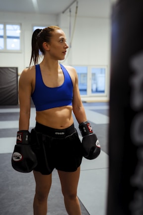 A female athlete in a gym setting wearing boxing gloves, a blue sports bra, and black shorts stands with an expression of focus and determination. The background features padded walls and large windows letting in natural light.