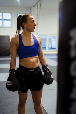 A female athlete in a gym setting wearing boxing gloves, a blue sports bra, and black shorts stands with an expression of focus and determination. The background features padded walls and large windows letting in natural light.