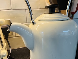 A white kettle with a chrome handle sits on a kitchen counter. The background includes a tiled backsplash and a nearby plug socket. The lid of the kettle has a black top and the design is simple and classic.