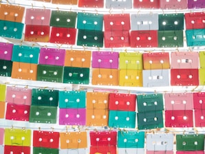 An array of colorful cassette tapes is displayed in a grid pattern, neatly arranged by rows and attached with small hooks. The tapes feature vibrant colors such as pink, green, red, orange, yellow, and teal, creating a visually striking and retro-inspired aesthetic. The overall composition is artistic and creates a sense of nostalgia.