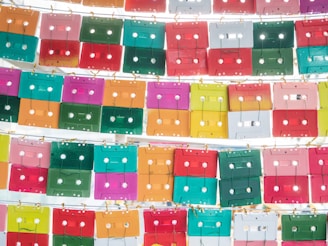 An array of colorful cassette tapes is displayed in a grid pattern, neatly arranged by rows and attached with small hooks. The tapes feature vibrant colors such as pink, green, red, orange, yellow, and teal, creating a visually striking and retro-inspired aesthetic. The overall composition is artistic and creates a sense of nostalgia.