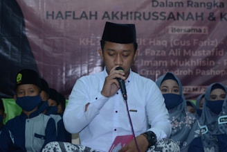 A group of people, mostly wearing traditional clothing and face masks, are present. The central figure is holding a microphone, possibly speaking or singing, and is dressed in a white shirt with a black cap. The background features a banner with text in another language.