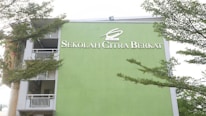 A building facade painted in light green with the words 'Sekolah Citra Berkat' in white lettering. The structure has several floors visible along with white railings and open balcony spaces. Lush green tree branches partially frame the building against a pale sky.