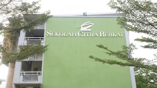 A building facade painted in light green with the words 'Sekolah Citra Berkat' in white lettering. The structure has several floors visible along with white railings and open balcony spaces. Lush green tree branches partially frame the building against a pale sky.