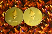 Two gold coins featuring the Ethereum logo are surrounded by numerous shiny, orange gemstones on a dark background. The coins have the Ethereum symbol and the word 'Ethereum' embossed on them.