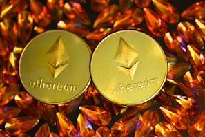 Two gold coins featuring the Ethereum logo are surrounded by numerous shiny, orange gemstones on a dark background. The coins have the Ethereum symbol and the word 'Ethereum' embossed on them.