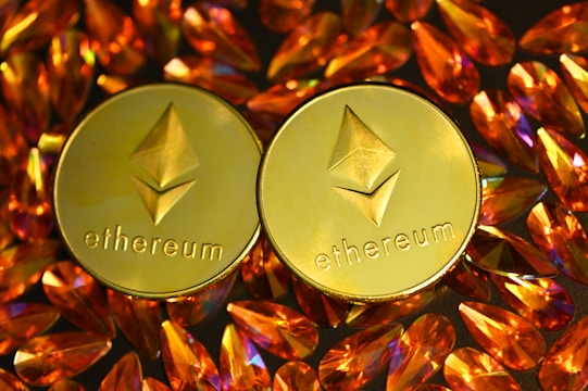 Two gold coins featuring the Ethereum logo are surrounded by numerous shiny, orange gemstones on a dark background. The coins have the Ethereum symbol and the word 'Ethereum' embossed on them.