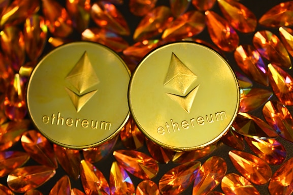 Two gold coins featuring the Ethereum logo are surrounded by numerous shiny, orange gemstones on a dark background. The coins have the Ethereum symbol and the word 'Ethereum' embossed on them.