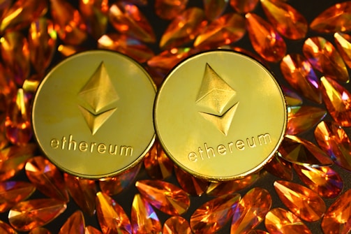 Two gold coins featuring the Ethereum logo are surrounded by numerous shiny, orange gemstones on a dark background. The coins have the Ethereum symbol and the word 'Ethereum' embossed on them.