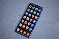 A smartphone with a large screen displaying various colorful app icons arranged in a grid. The smartphone is laying on a gray, speckled surface.