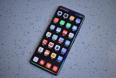 A smartphone with a large screen displaying various colorful app icons arranged in a grid. The smartphone is laying on a gray, speckled surface.