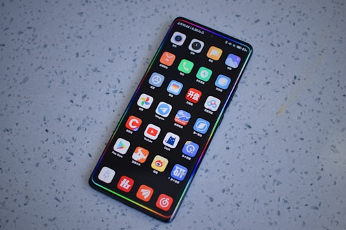 A smartphone with a large screen displaying various colorful app icons arranged in a grid. The smartphone is laying on a gray, speckled surface.
