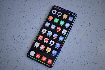 A smartphone with a large screen displaying various colorful app icons arranged in a grid. The smartphone is laying on a gray, speckled surface.