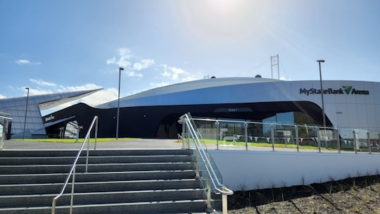 A modern architectural structure with sleek, curved lines and a combination of black and white surfaces. The MyState Bank Arena signage is prominently displayed on the building. The foreground features stairs leading up to the entrance, with metal railings and a glass barrier. The sky is clear and sunny, casting shadows across the building.