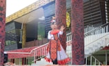 A child is dressed in traditional clothing, performing a cultural dance. The outfit is decorated with intricate patterns and vibrant colors, predominantly red and white. The background includes ornately designed pillars and a building with a patterned facade.
