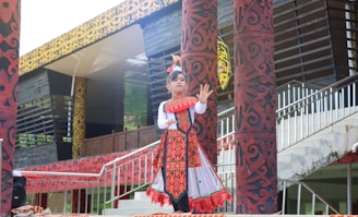 A child is dressed in traditional clothing, performing a cultural dance. The outfit is decorated with intricate patterns and vibrant colors, predominantly red and white. The background includes ornately designed pillars and a building with a patterned facade.