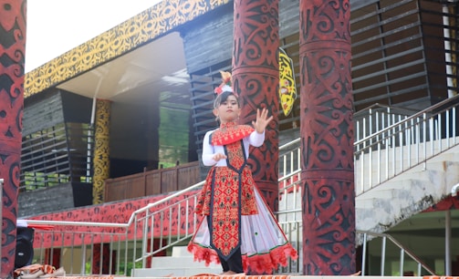 A child is dressed in traditional clothing, performing a cultural dance. The outfit is decorated with intricate patterns and vibrant colors, predominantly red and white. The background includes ornately designed pillars and a building with a patterned facade.